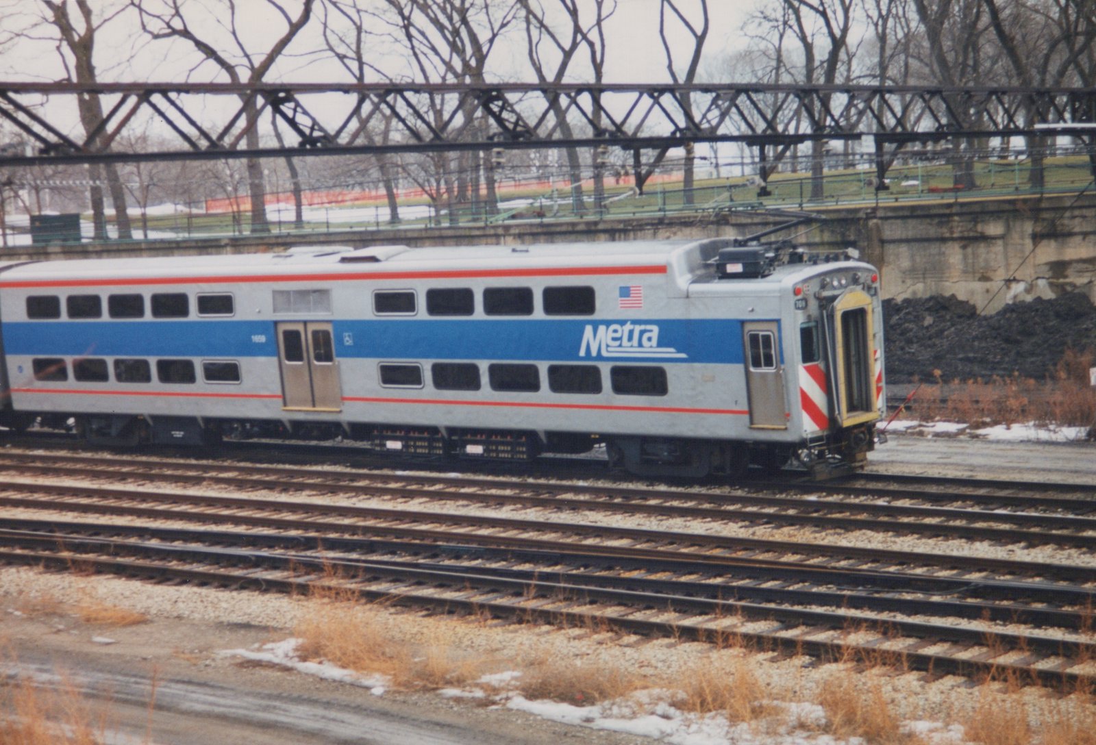 A silver, blue, and red double-decker Metra train car with the number 1659 sits on tracks under a bridge, with snow on the ground.