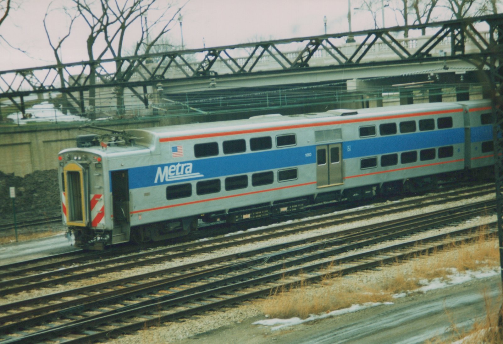 Two silver, blue, and red Metra Highliner train cars, numbered 1660, are on railroad tracks under a steel bridge, with snow on the ground and bare trees in the background.