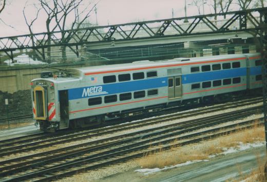 Two silver, blue, and red Metra Highliner train cars, numbered 1660, are on railroad tracks under a steel bridge, with snow on the ground and bare trees in the background.