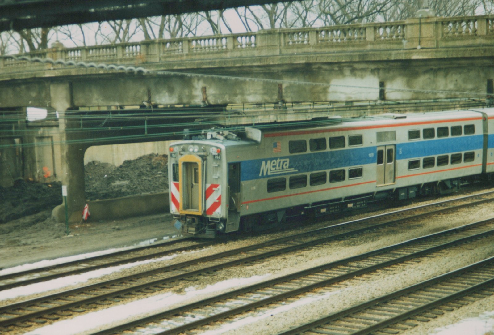 A Metra commuter train car, number 1578, painted silver with blue and red stripes, is stopped on tracks under a concrete bridge with snow on the ground.