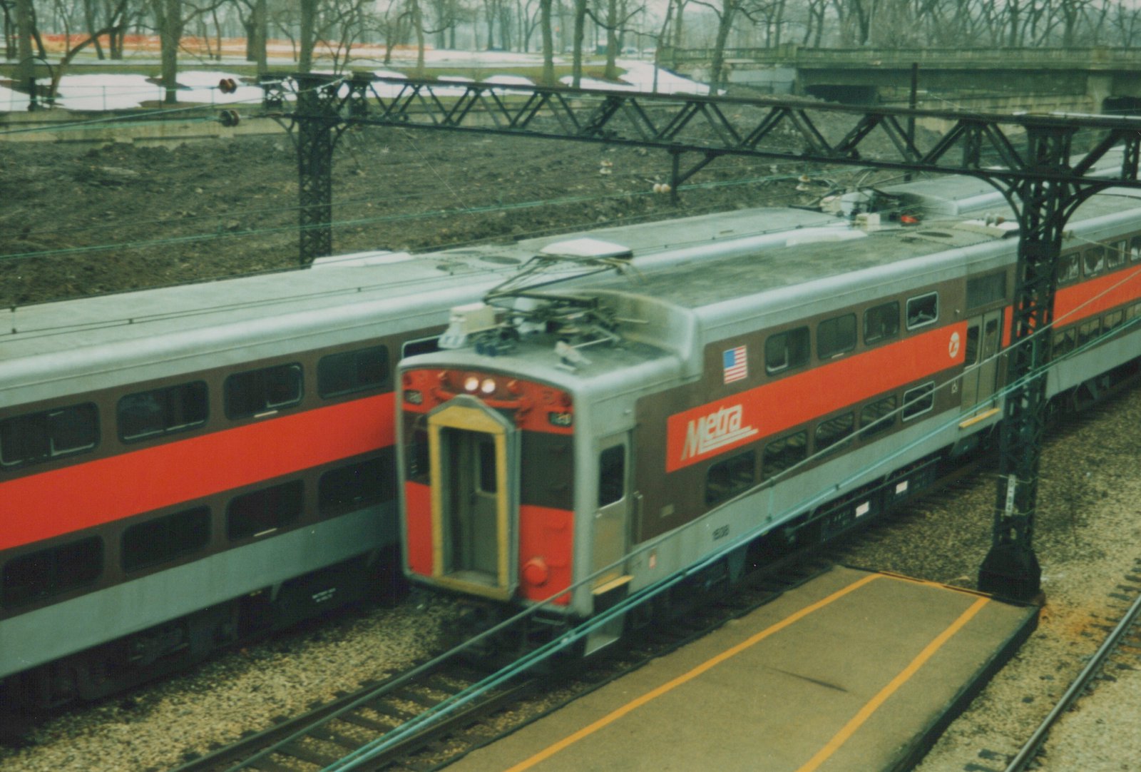 A silver, red, and brown Metra Highliner train car with the number 124, stopped at a station platform in a winter scene with snow on the ground and bare trees in the background.