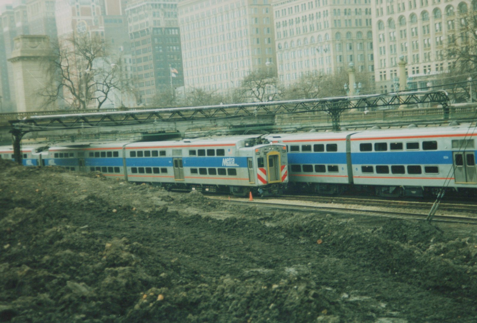 A silver, blue, and red Metra train with the number 1634 is shown on tracks next to a large pile of dirt, with tall city buildings and an elevated railway structure behind it.