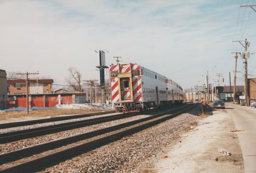 A silver double-decker commuter train with red and white striped ends moves along tracks bordered by snow and utility poles under a clear sky.