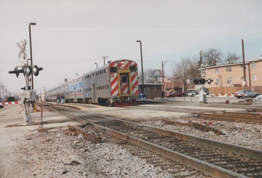 A long, silver Metra commuter train with red and white striped cab ends is stopped at a railroad crossing. The ground is bare and patchy with snow, and a few people stand on the platform. A grade crossing sign shows three tracks.