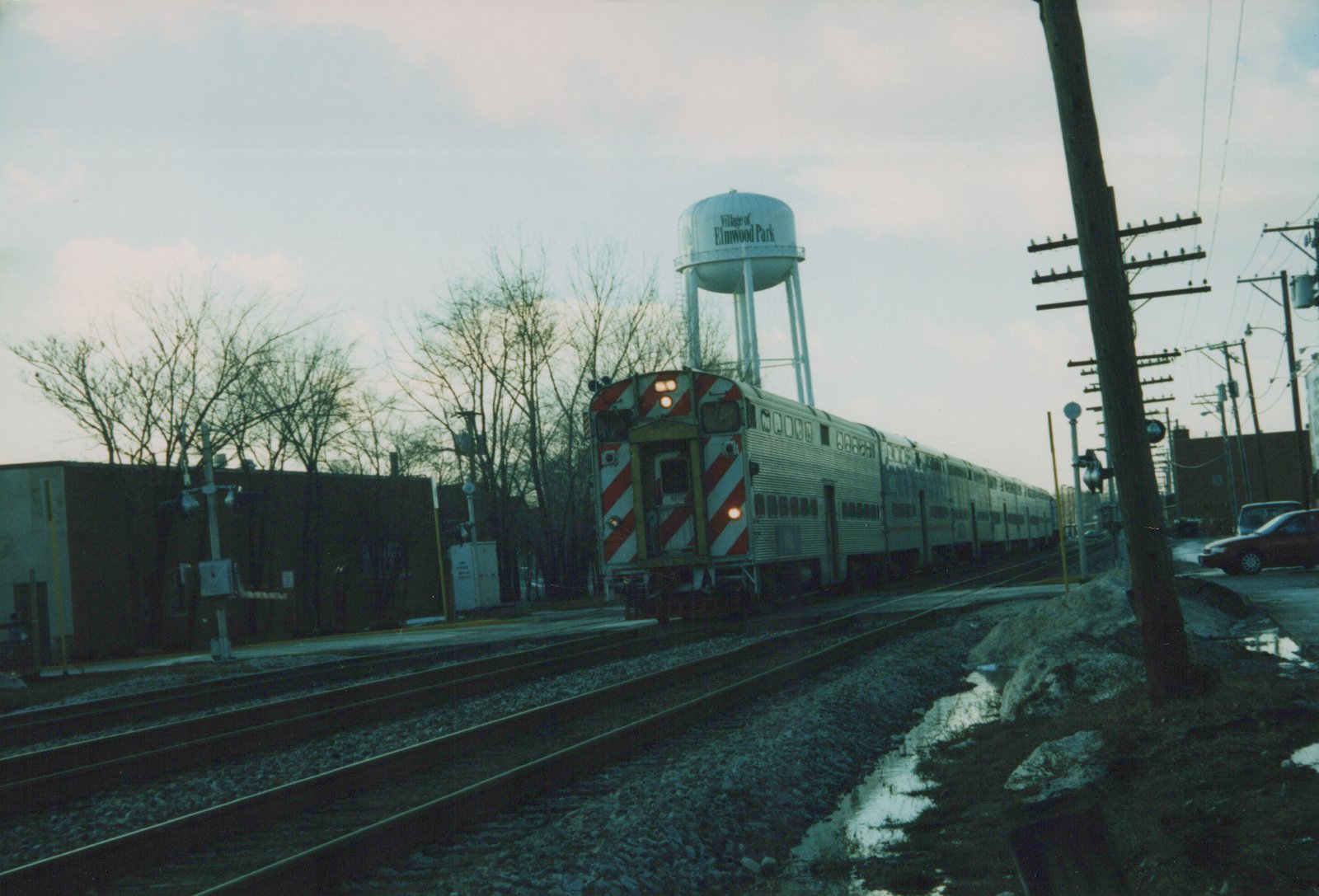 A silver Metra Gallery commuter train with red and white striped rear end is stopped at a station with a water tower in the background in Elmwood Park, Illinois.