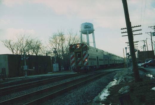 A silver Metra Gallery commuter train with red and white striped rear end is stopped at a station with a water tower in the background in Elmwood Park, Illinois.