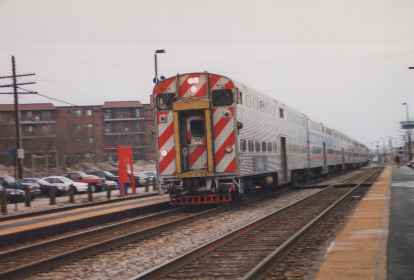 A Metra train with a silver and red-striped locomotive numbered 8223 is seen from the rear, pushing several silver double-decker passenger cars along a set of tracks adjacent to a concrete station platform.