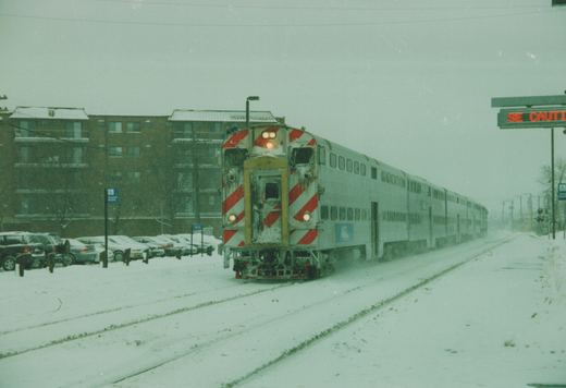 A silver Metra train with red and white striped cab ends moves along snow-covered tracks, with a brick building and parked cars in the background.