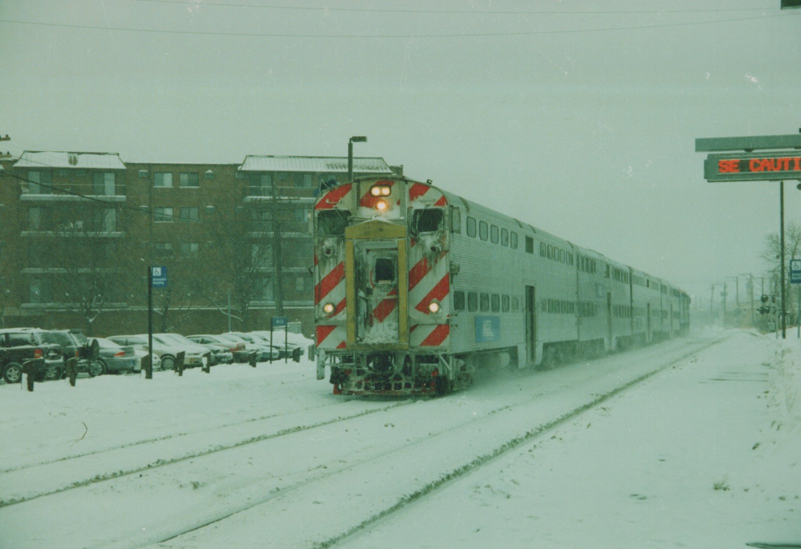 A silver Metra train with red and white striped cab ends moves along snow-covered tracks, with a brick building and parked cars in the background.