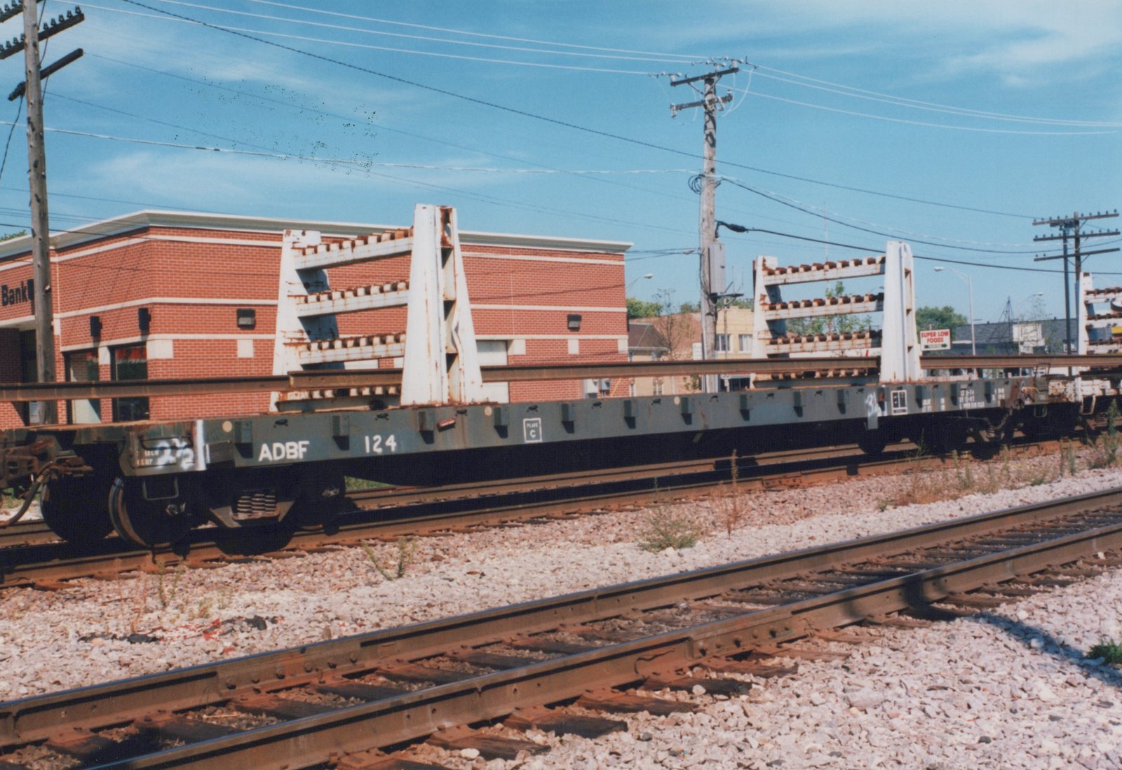 A side view of a freight car labeled ADBF 124 carrying long steel rails on a railroad track, with a brick building and power lines in the background under a clear blue sky.