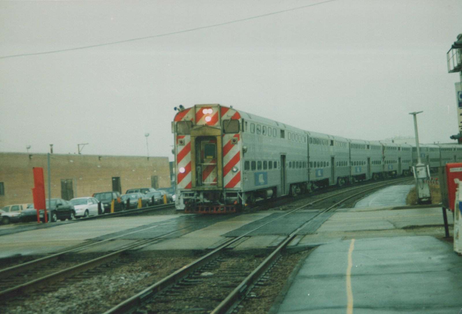 A silver double-decker Metra commuter train with red-and-white striped ends sits on tracks at a grade crossing, with parked cars and a brick building in the background under an overcast sky.
