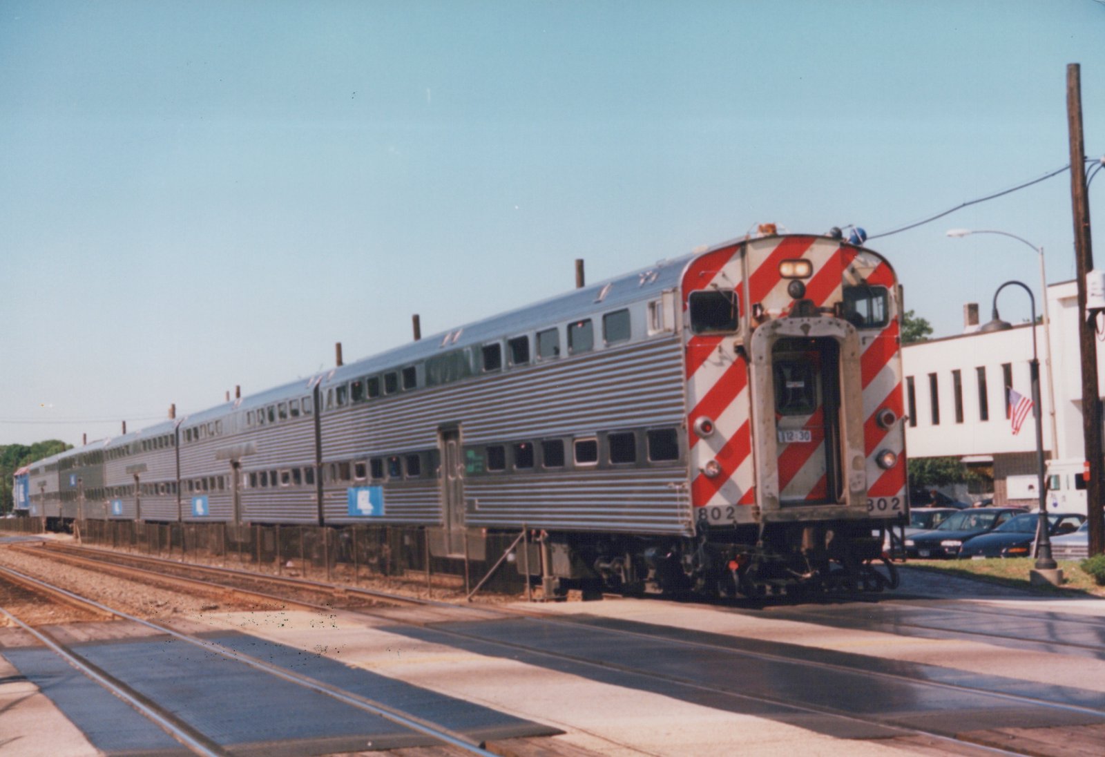A long, double-decker stainless steel Metra commuter train is stopped at a street-level crossing, with its rear end facing the camera and showing the fleet number 802 and 302.