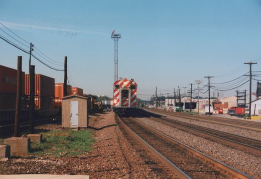 A red and white striped Metra cab car, number 804, viewed from the rear as it sits on tracks in a railroad yard with shipping containers and industrial buildings in the background.