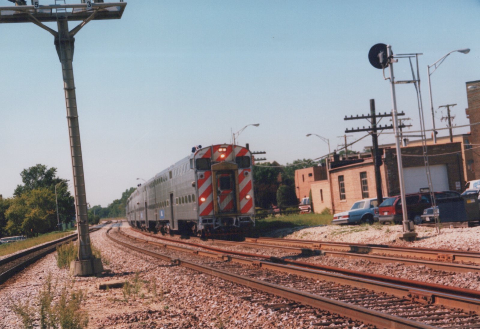 A silver Metra Highliner train with red diagonal stripes on its end travels along curving tracks under a clear blue sky, with parked cars and brick buildings in the background.