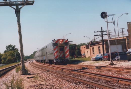 A silver Metra Highliner train with red diagonal stripes on its end travels along curving tracks under a clear blue sky, with parked cars and brick buildings in the background.