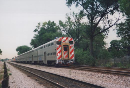 A silver Metra train with a red and white striped cab end is moving on tracks bordered by trees and gravel.