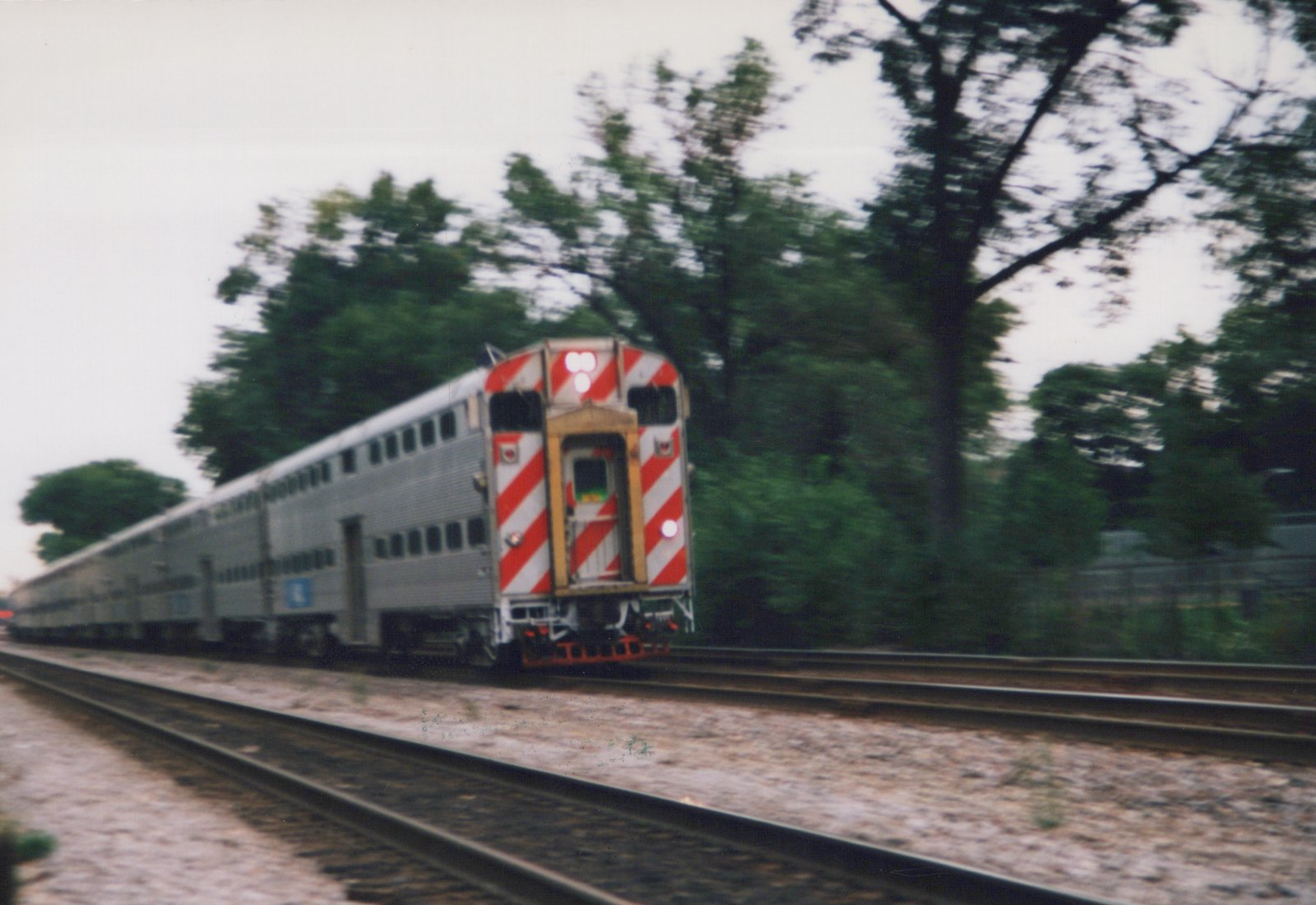 A silver Metra Gallery train with red and white striped ends is captured in motion on a tree-lined track on a cloudy summer day.