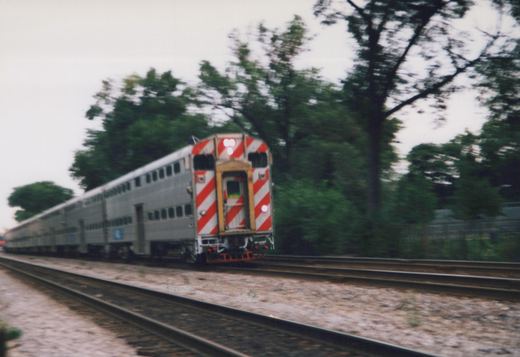 A silver Metra Gallery train with red and white striped ends is captured in motion on a tree-lined track on a cloudy summer day.