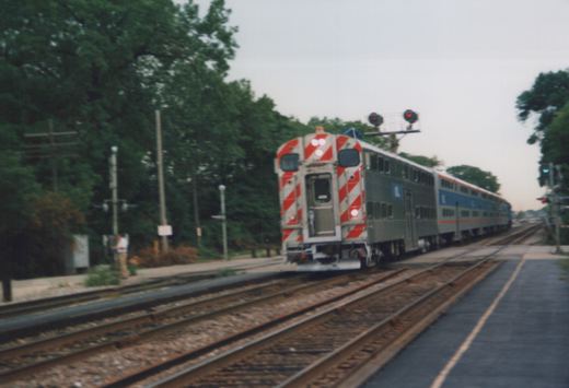 A silver Metra train with red and blue stripes, featuring the 'M' logo, travels on multiple tracks next to a station platform with a person standing on it, surrounded by trees on an overcast day.