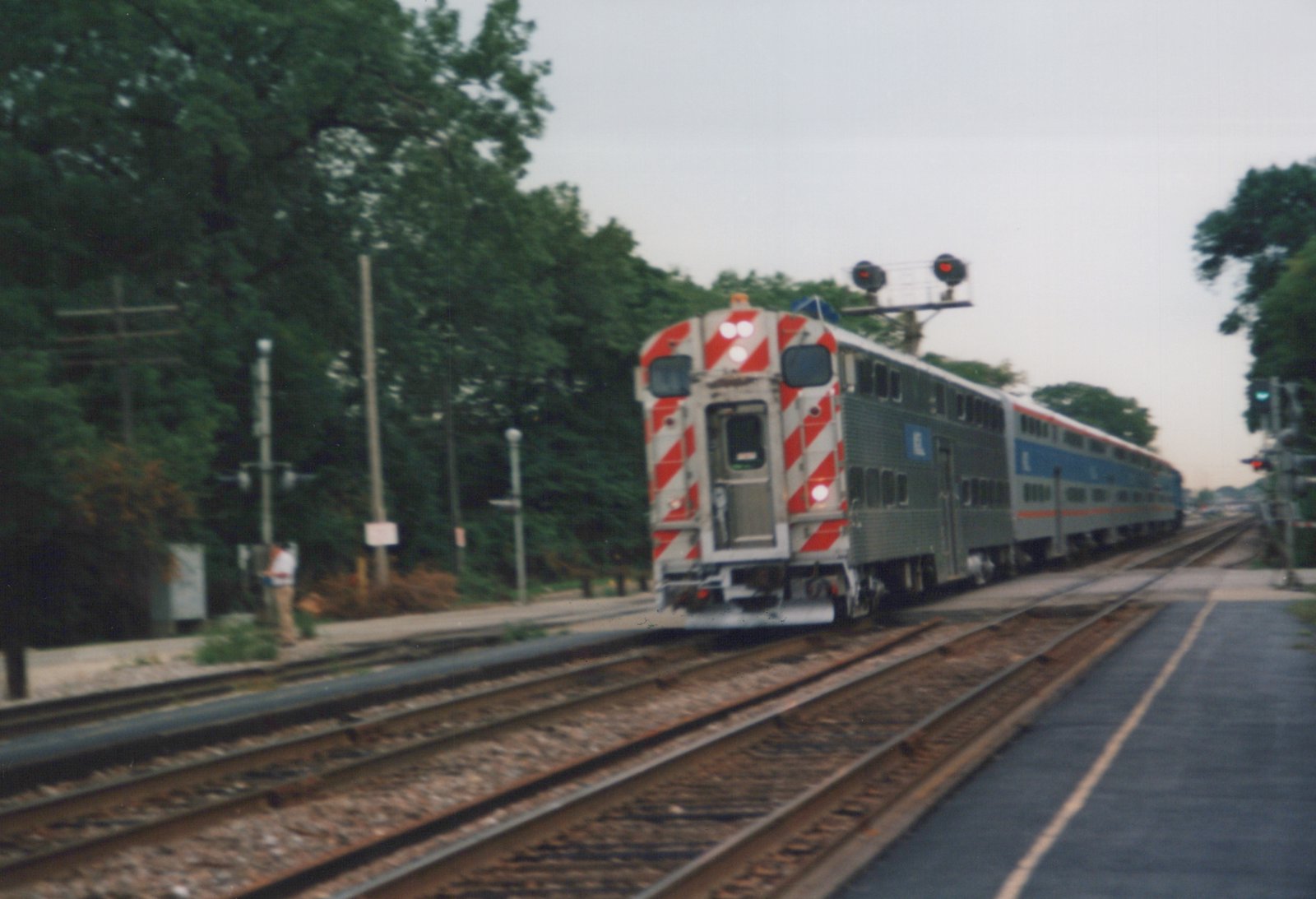 A silver Metra train with red and blue stripes, featuring the 'M' logo, travels on multiple tracks next to a station platform with a person standing on it, surrounded by trees on an overcast day.