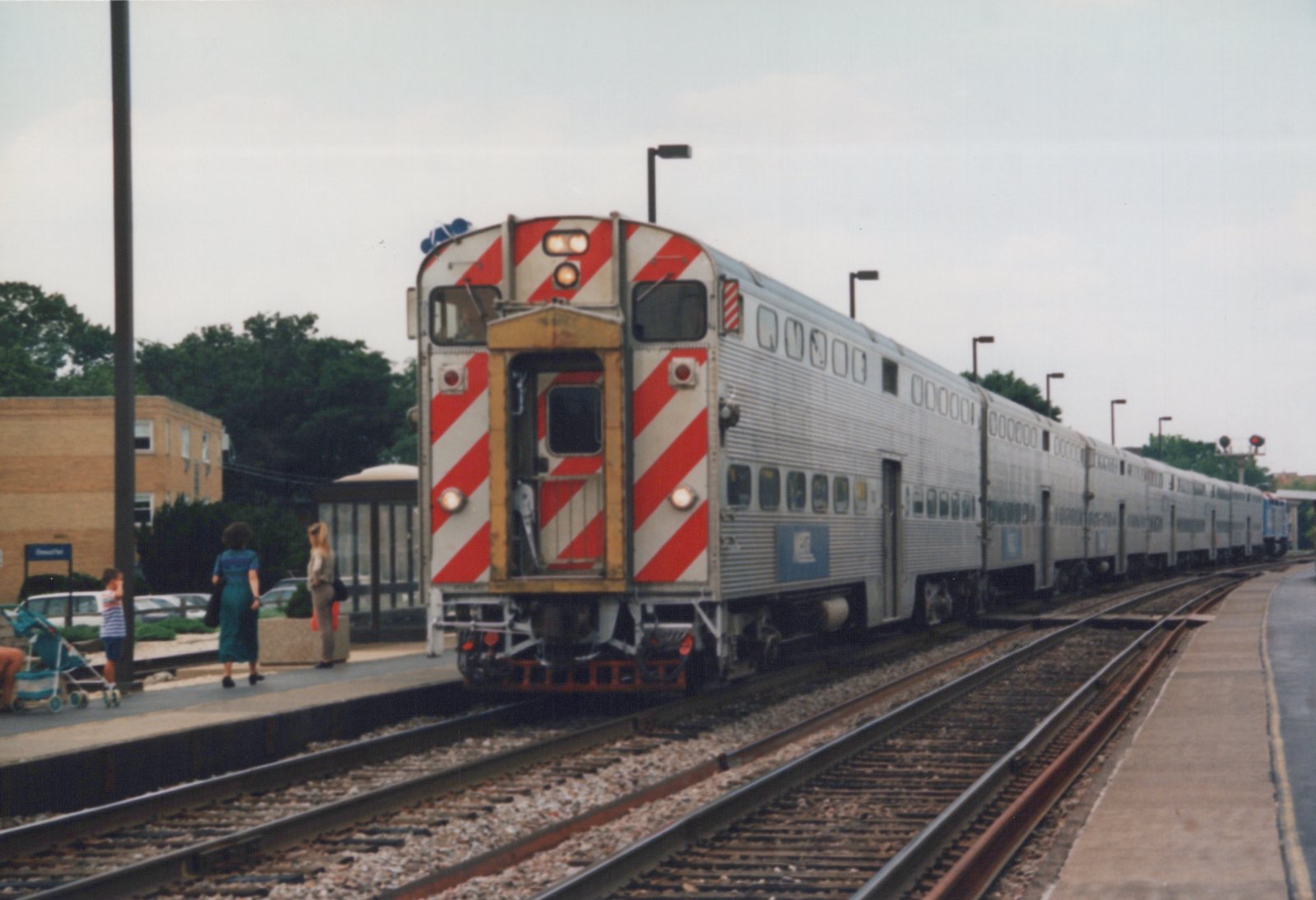 A silver, double-decker Metra commuter train with red and white striped cab end is stopped at a station platform where several people are waiting.