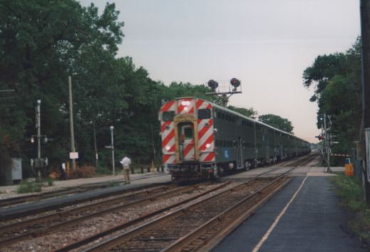 A Metra Highliner train, gray with a blue stripe and logo, travels on tracks with a red-and-white striped rear end, next to a station platform with two people standing on it.