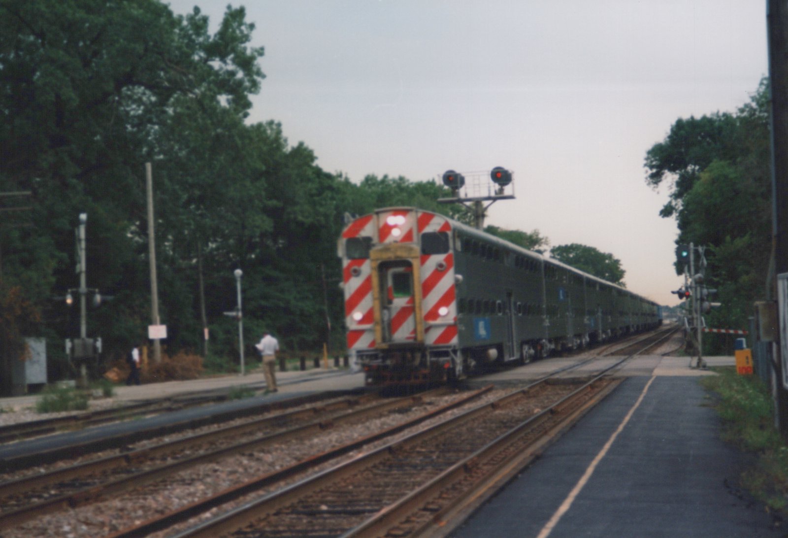 A Metra Highliner train, gray with a blue stripe and logo, travels on tracks with a red-and-white striped rear end, next to a station platform with two people standing on it.