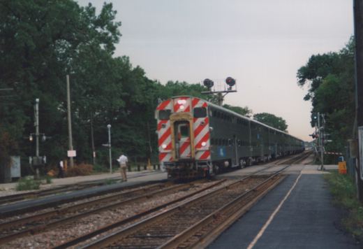 A gray Budd RDC railcar with red-and-white striped front, marked with the Chicago and North Western logo, is seen from a three-quarter rear view as it travels on tracks beside a platform, with trees and a grade crossing signal in the background under an overcast sky.