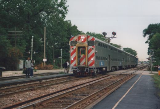 A silver Metra commuter train with red and white striped ends is stopped at a small station platform with several people waiting; trees line the background and railroad tracks extend into the distance.