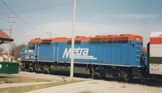 Side view of a blue and orange Metra diesel locomotive number 162 on a track next to a sidewalk and utility pole, with a commuter car partially visible behind it.