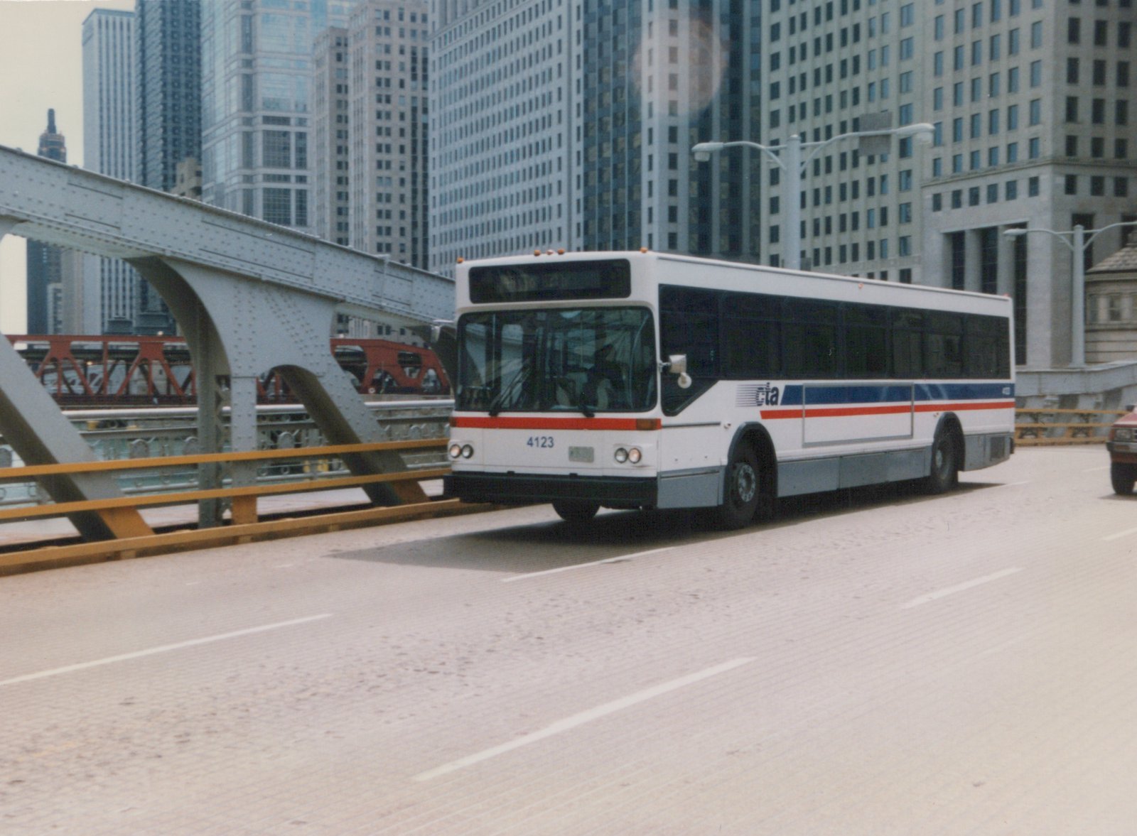 A white, red, and blue Chicago Transit Authority bus, number 4123, is driving on a bridge in a city with tall buildings in the background.