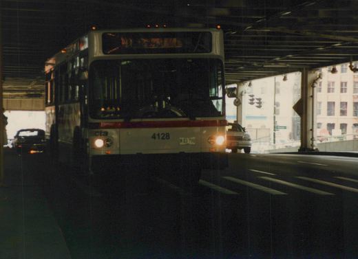 Front view of a white and red Chicago Transit Authority motor bus, number 4128, with headlights on, driving under an overpass on a city street.