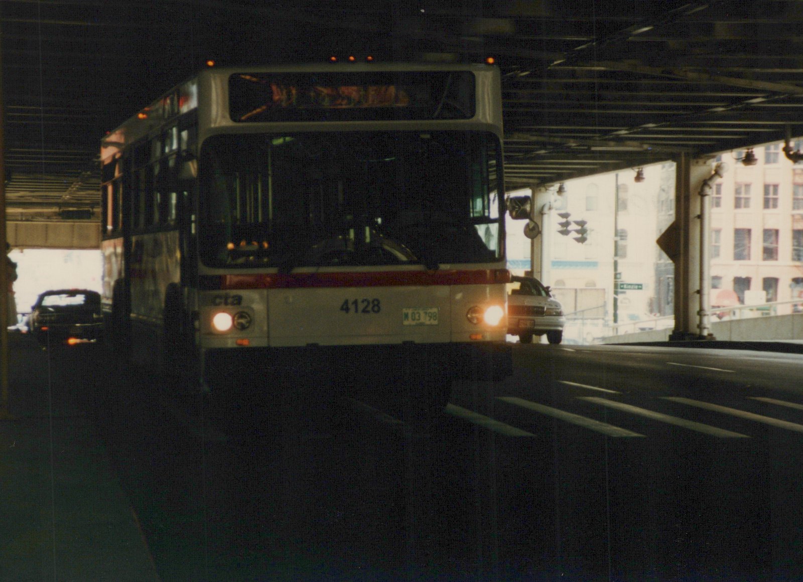 Front view of a white and red Chicago Transit Authority motor bus, number 4128, with headlights on, driving under an overpass on a city street.
