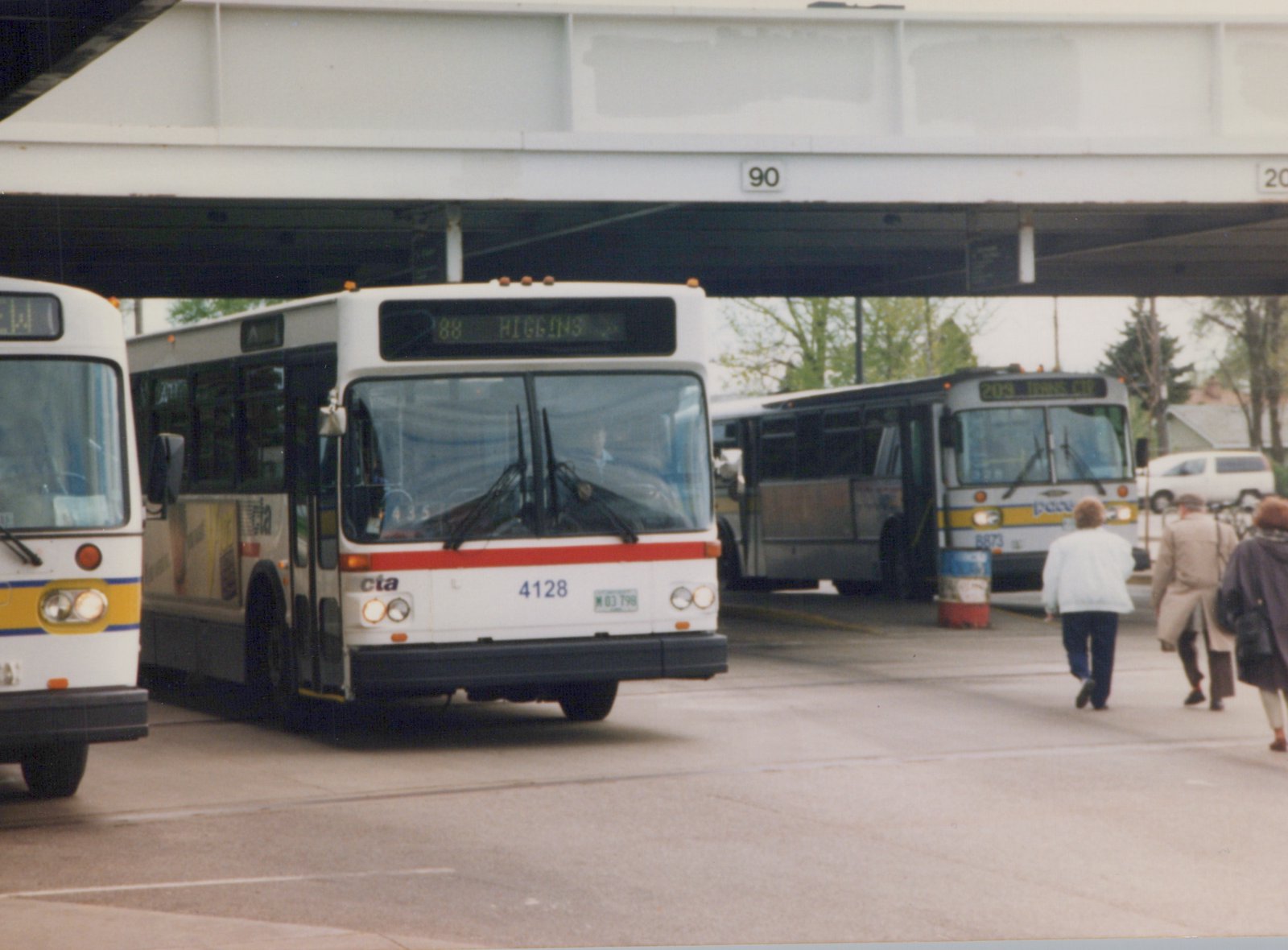 Front view of a white Chicago Transit Authority bus with a red stripe and the number 4128, next to a white Pace bus with a yellow and blue stripe, under a steel overpass with the number 90, with three people walking nearby.