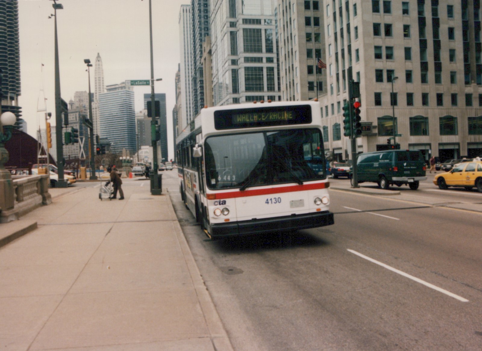 Front view of a white and red CTA bus numbered 4130 with 'WALLACE/RACINE' on its destination sign, driving on a city street with tall buildings and other vehicles in the background.