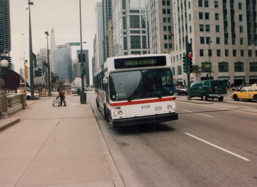 Front view of a white and red CTA bus numbered 4130 with 'WALLACE/RACINE' on its destination sign, driving on a city street with tall buildings and other vehicles in the background.