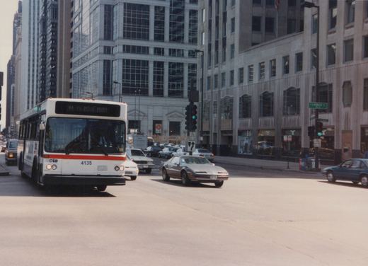 A white and red Chicago Transit Authority bus, number 4135, drives on a city street with other cars and tall buildings in the background.