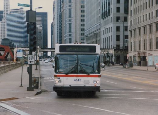 Front view of a white and red Chicago Transit Authority bus, number 4143, driving on a city street with tall buildings and a bridge in the background.