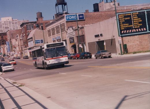 A white Chicago Transit Authority bus with red and blue stripes, number 4149, is driving down a wide city street lined with brick buildings and parked cars under a clear blue sky.
