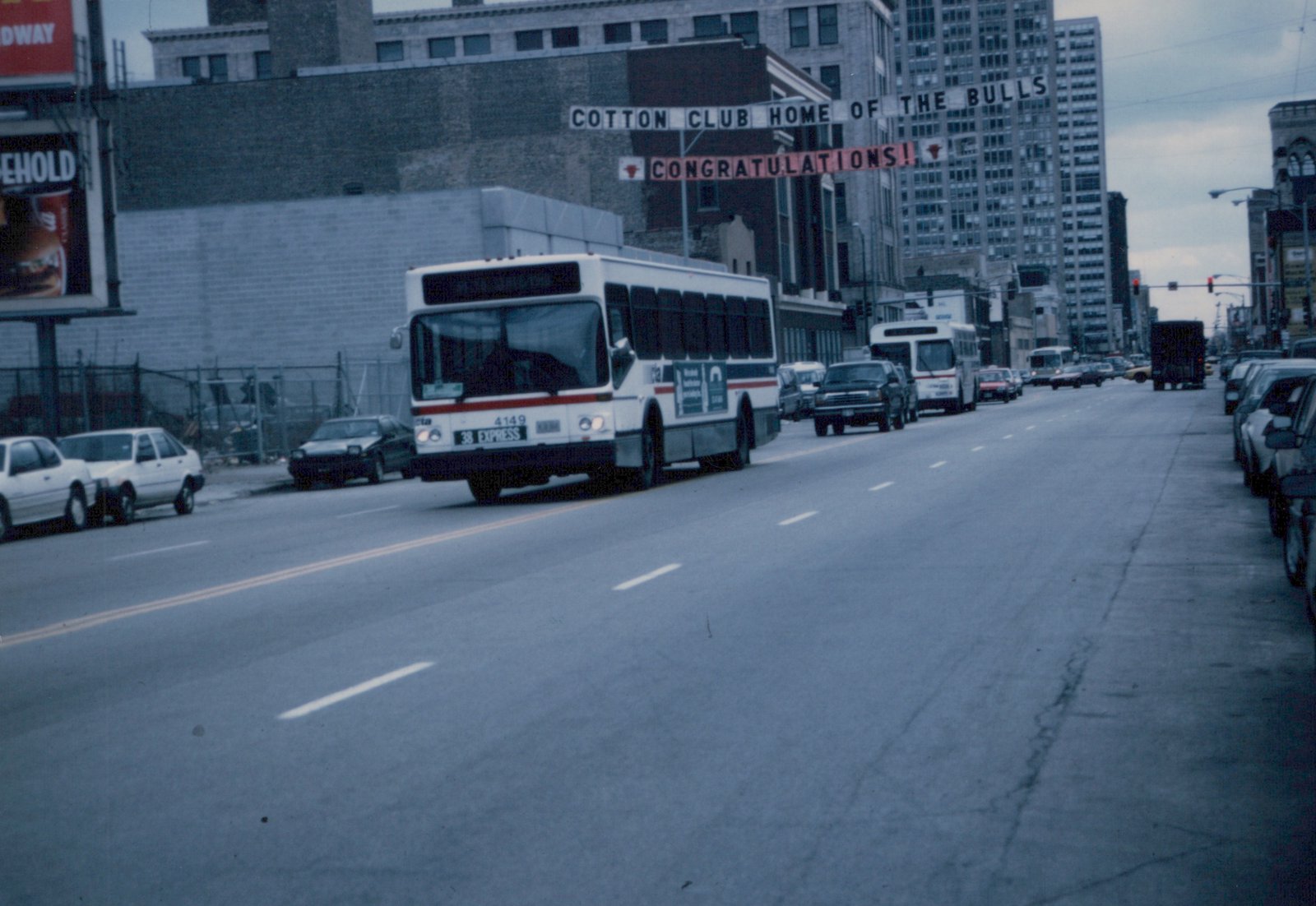 A white CTA bus with the number 4149 is driving down a city street with other cars and a large sign for the Cotton Club and the Chicago Bulls in the background.