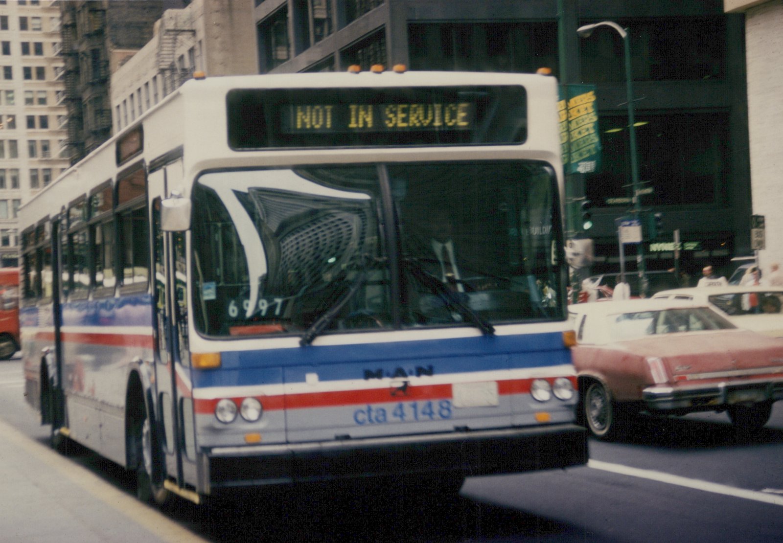 A white, blue, and red Chicago Transit Authority bus with the number 4148 on the front bumper and 'NOT IN SERVICE' on the destination sign, driving on a city street with a sedan alongside it.