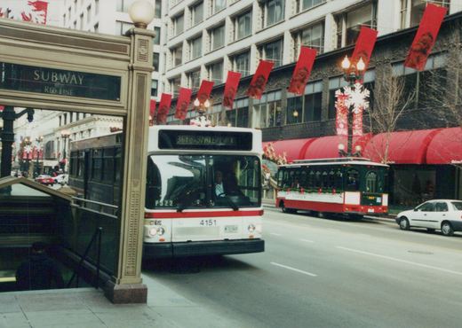 A white and red CTA bus with the number 4151 and a green and red trolleybus with the number 397 are on a city street decorated for Christmas, with a subway entrance to the left.