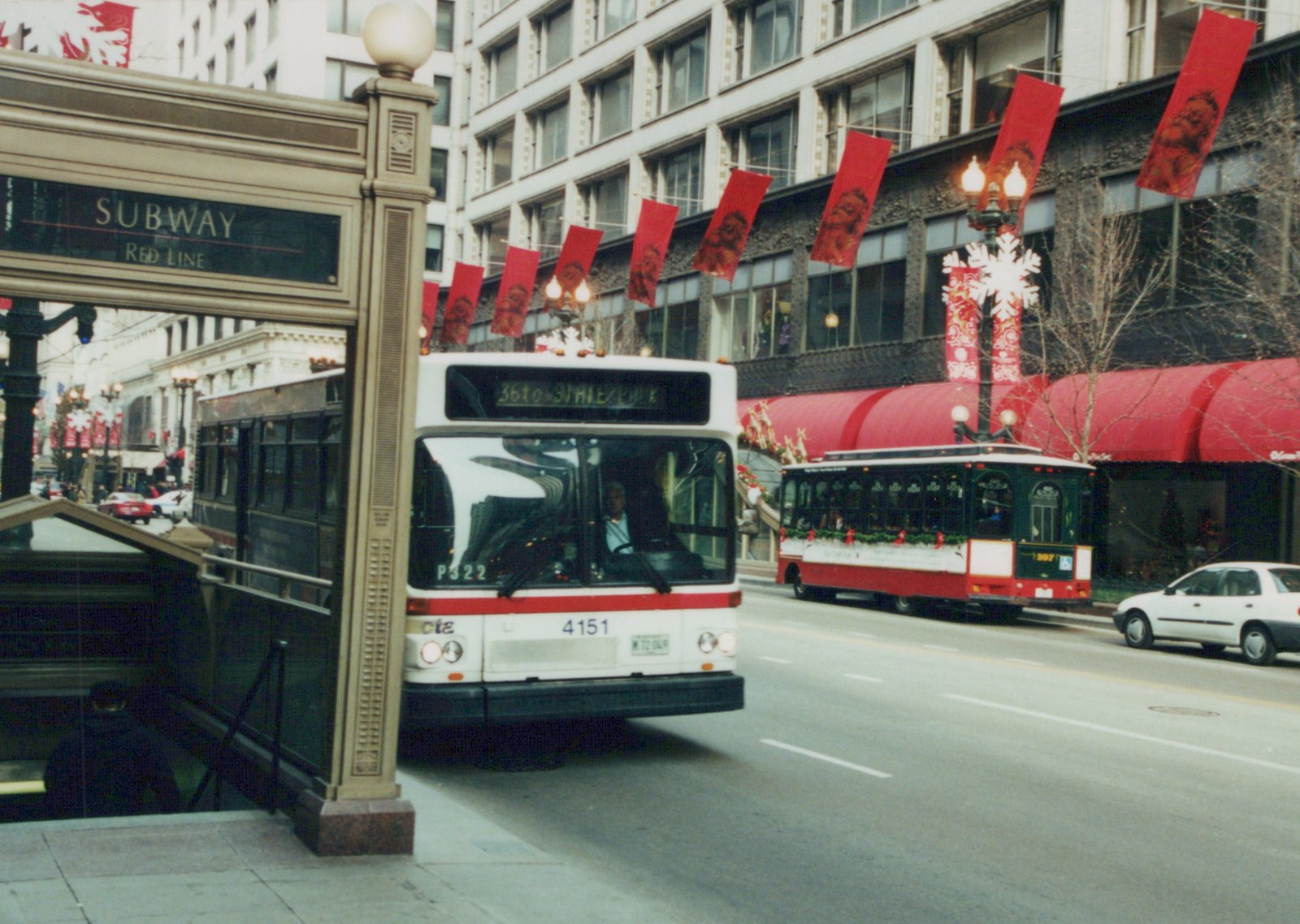 A white and red CTA bus with the number 4151 and a green and red trolleybus with the number 397 are on a city street decorated for Christmas, with a subway entrance to the left.