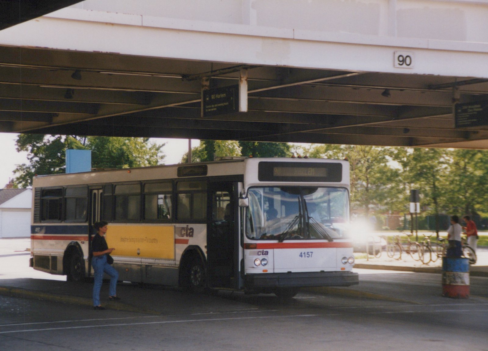 A white and blue CTA bus with number 4157 is stopped under an elevated structure. A passenger is boarding, and bicycles are parked nearby. The destination sign reads '90 Harlem'.