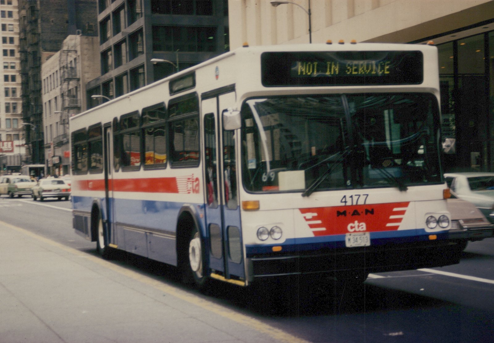 A white and blue Chicago Transit Authority bus with a red stripe, numbered 4177 and displaying 'NOT IN SERVICE' on its front sign, is moving down a street with buildings and other cars in the background.