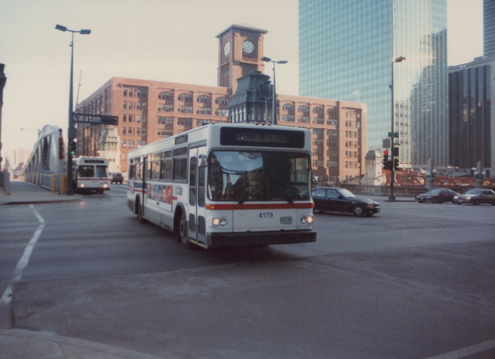 A white and red Chicago Transit Authority bus, number 4179, is driving on a city street with another CTA bus and cars in the background, near a historic clock tower and tall glass buildings.