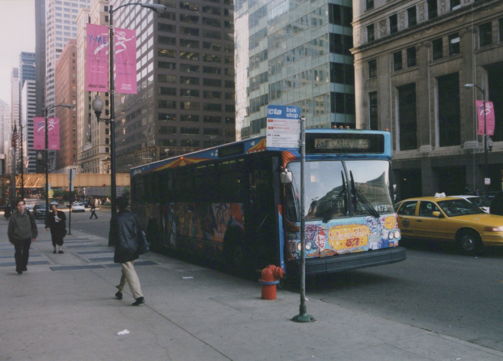 A blue and art-covered CTA bus numbered 4175 is stopped on a Chicago street with pedestrians walking on the sidewalk and a yellow taxi cab nearby.