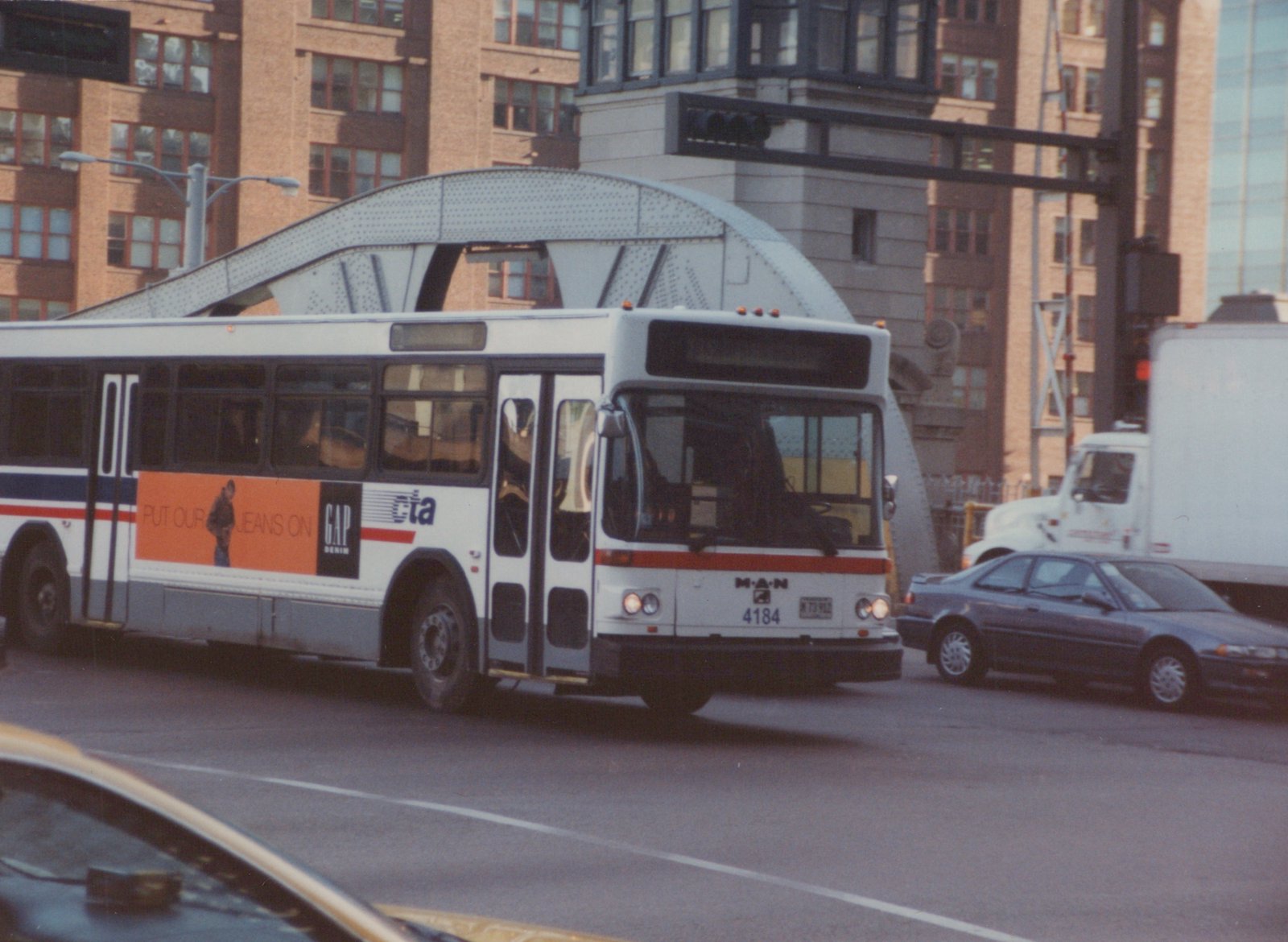 A white and blue Chicago Transit Authority bus with the number 4184 is driving on a city street under a metal arch bridge, with other cars and tall buildings in the background.