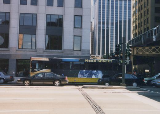 A side view of a dark blue diesel bus with a space-themed advertisement driving on a city street, with cars, modern buildings, and an elevated train visible.