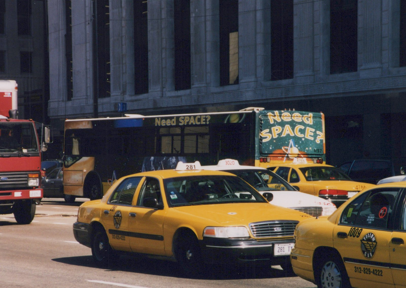 A dark green Chicago Transit Authority bus with the number 4187 on its rear, surrounded by yellow taxis and a red truck on a city street on a sunny day.